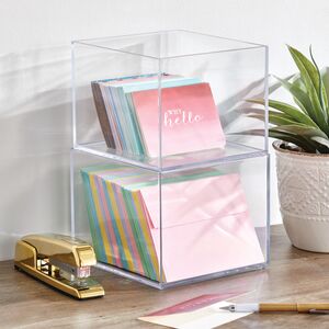 clear stacked bins holding note cards, envelopes, gold stapler, plant on wood shelf, white wall