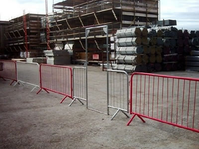 Galvanized and red powder coated crowd control barriers and a crowd control barrier gate are standing on the concrete in factory.